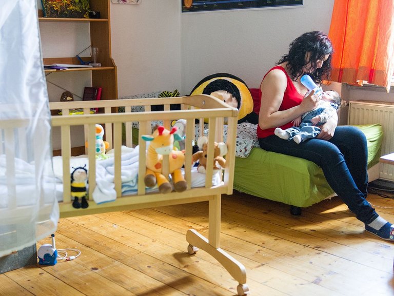 Eine Mutter füttert ihren Säugling mit der Flasche und sitzt dabei auf einem grünen Bett.