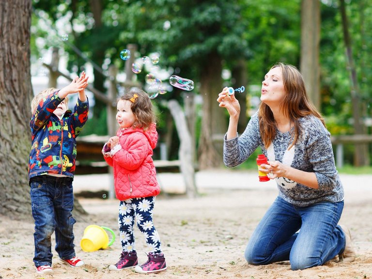 Zwei Kinder spielen mit ihrer Mama draußen und fangen Seifenblasen mit ihren Händen.