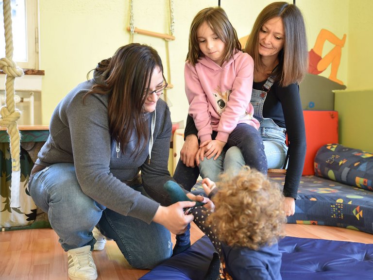 Eine Mutter Zieht ihrem Kind eine Socke an und ein anderes Kind sitzt auf dem Schoß von ihrer Mama und schaut zu.