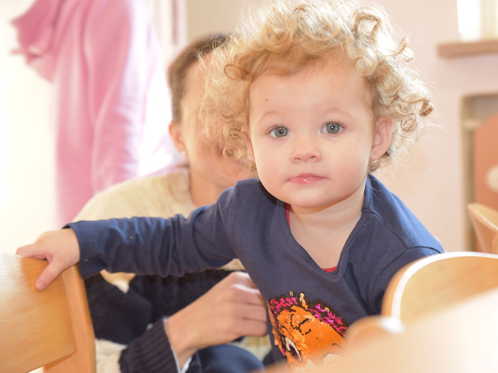 Kleines Kind mit lockigen Haaren sitzt auf dem Schoss von der Mutter und schaut in die Kamera