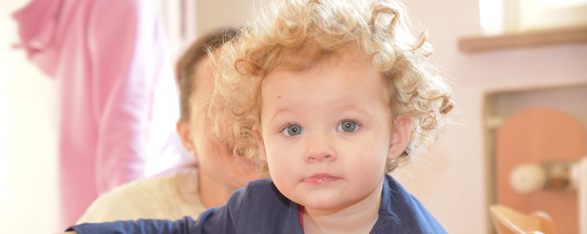 Kleines Kind mit lockigen Haaren sitzt auf dem Schoss von der Mutter und schaut in die Kamera